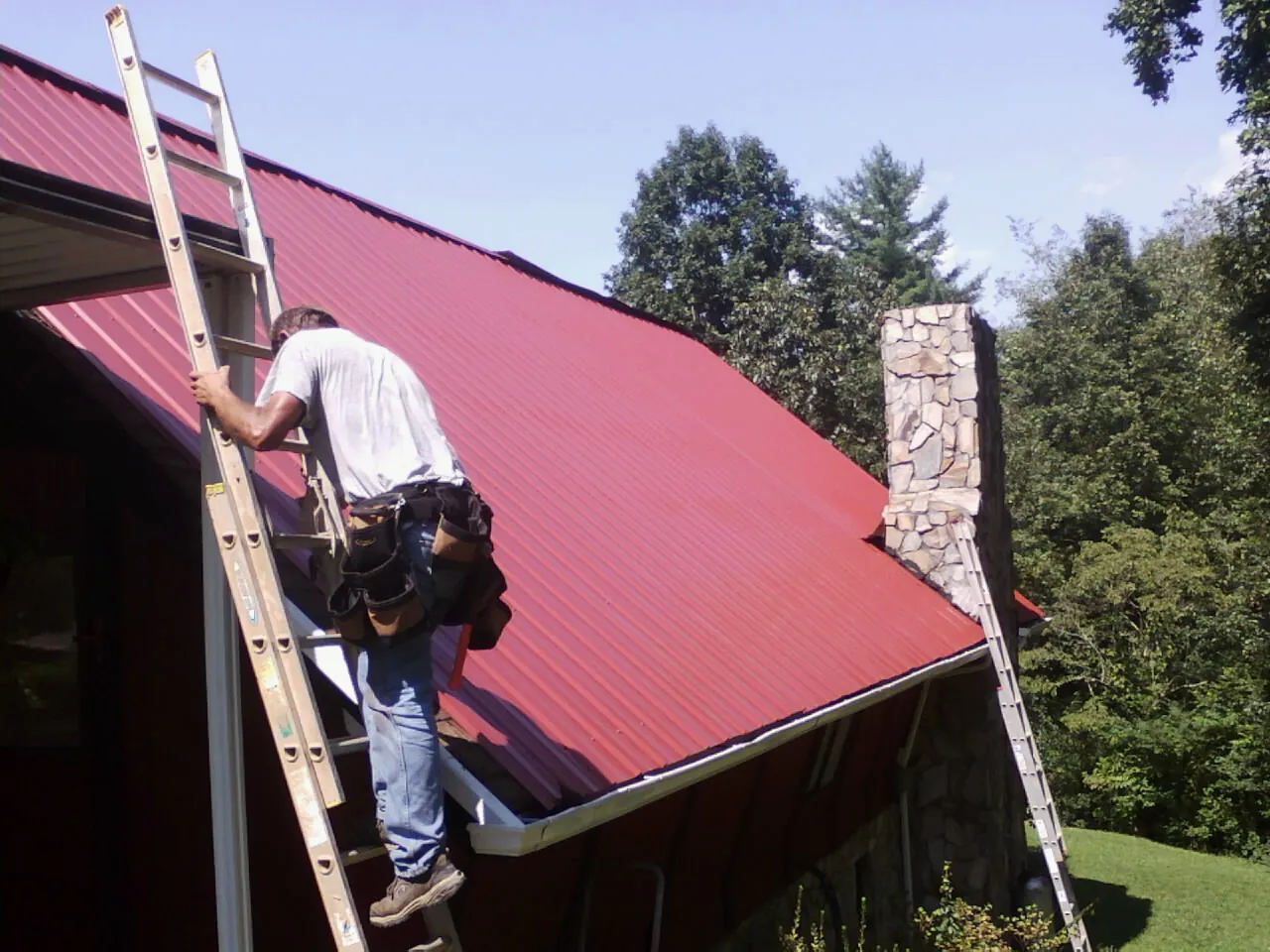 Roofer on a ladder beside a red metal roof and stone chimney — Soffit & Fascia Repair by Roofing Brothers in Jamieson